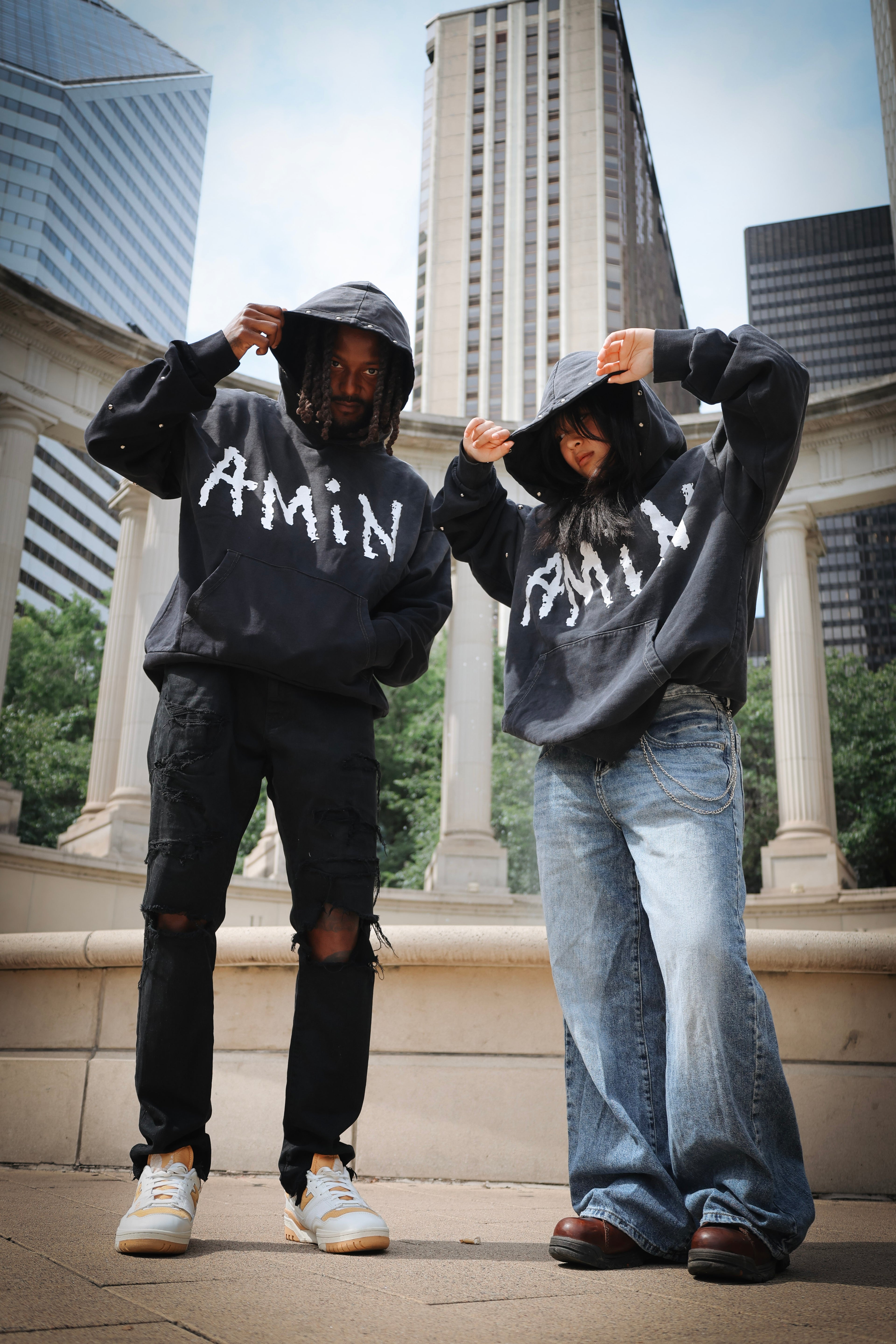 Two models wearing matching black hoodies with 'Amin' written in white across the chest, standing in front of a classical stone colonnade with skyscrapers in the background.